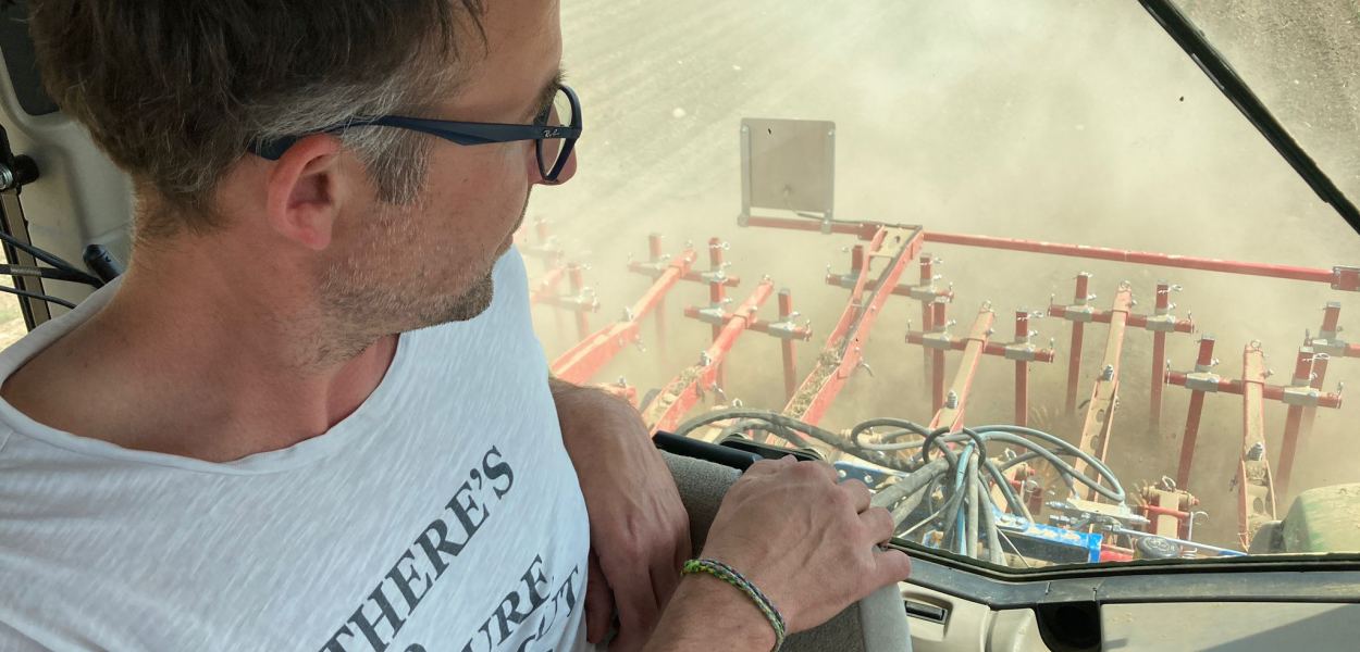 Soja-Feld mit Mann rechts im Bild, der in die Erde mit noch sehr kleinen Sojapflanzen greift. Im Hintergrund links in weiter Entfernung steht ein Traktor. Der Himmel ist blau, durchzogen von weißen Wolken. Foto: Althea Müller