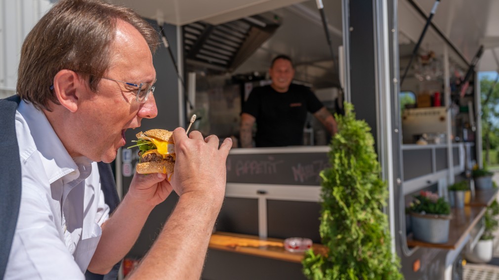 Bürgermeister Matthias Stadler beim Abbeißen von einem Burger, im Hintergrund sind der Foodtruck s'Hutsch und Thomas Haiderer zu sehen. (Foto: Arman Kalteis)