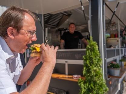 Bürgermeister Matthias Stadler beim Abbeißen von einem Burger, im Hintergrund sind der Foodtruck s'Hutsch und Thomas Haiderer zu sehen. (Foto: Arman Kalteis)