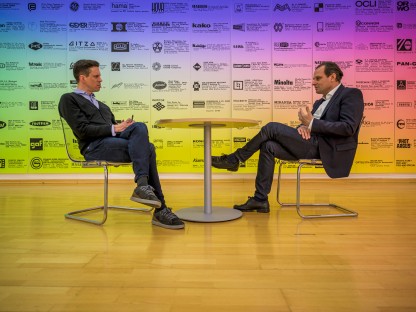 Michael Koppensteiner im Gespräch mit Alfred Kellner, sitzend, dazwischen ein runder Tisch, im Hintergrund eine farbig gestaltete Plakatwand. (Foto: Arman Kalteis)