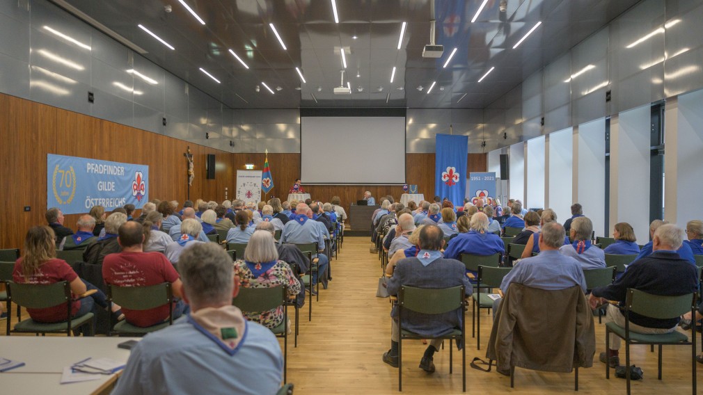 Viele Personen sittzen in einem Veranstaltungsraum und ören einem Redner zu.