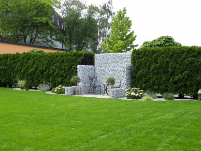 Blick auf Rasen in einen Garten im Hintergrund, eine Hecke, Sträucher und  Platz mit Steinmauer kleinen Tisch und zwei Sesseln. (Foto: Andrea Heinzl)