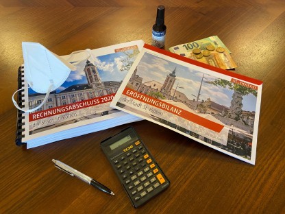Die Broschüren zum Rechnungsabschluss, ein Taschenrechner, Geldscheine und Münzen, eine Schutzmaske und ein Fläschchen Desinfektionsmittel auf einem Tisch. (Foto: Thomas Kainz)