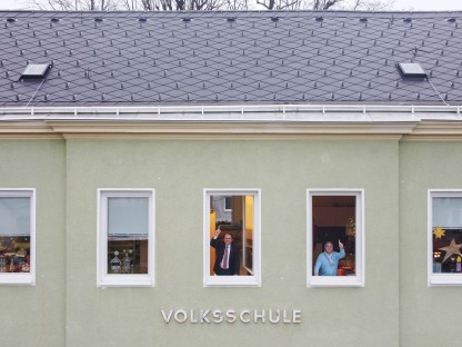 Bürgermeister Matthias Stadler und Schulamtsleiter Andreas Schmidt schauen aus Fenstern des Kindergarten- und Volksschul-Gebäude in Ratzersdorf. und zeigen nach oben in Richtung des Daches.