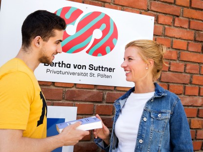 Ein Mann der einer Frau ein Buch reicht, vor einer Fassade mit dem Logo der Bertha von Suttner Privatuniversität St. Pölten. (Foto: Christian-Ariel-Heredia)