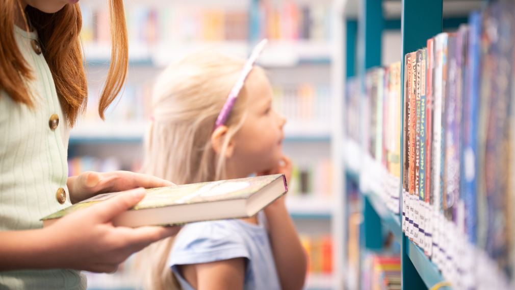 Ein Buch in der Hand eines Mädchens, verschwommen im Hintergrund schmökert ein jüngeres Mädchen durch die Bücher der Bücherei.