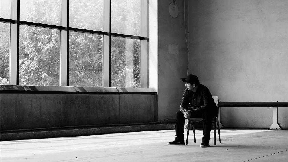 Schwarz-Weiß Foto des Musikers CHiLL-iLL. Er sitzt in einem großen Raum auf einem Stuhl und schaut durch eine große Fensterfront in die Natur hinaus. (Foto: Fabian Fallend)