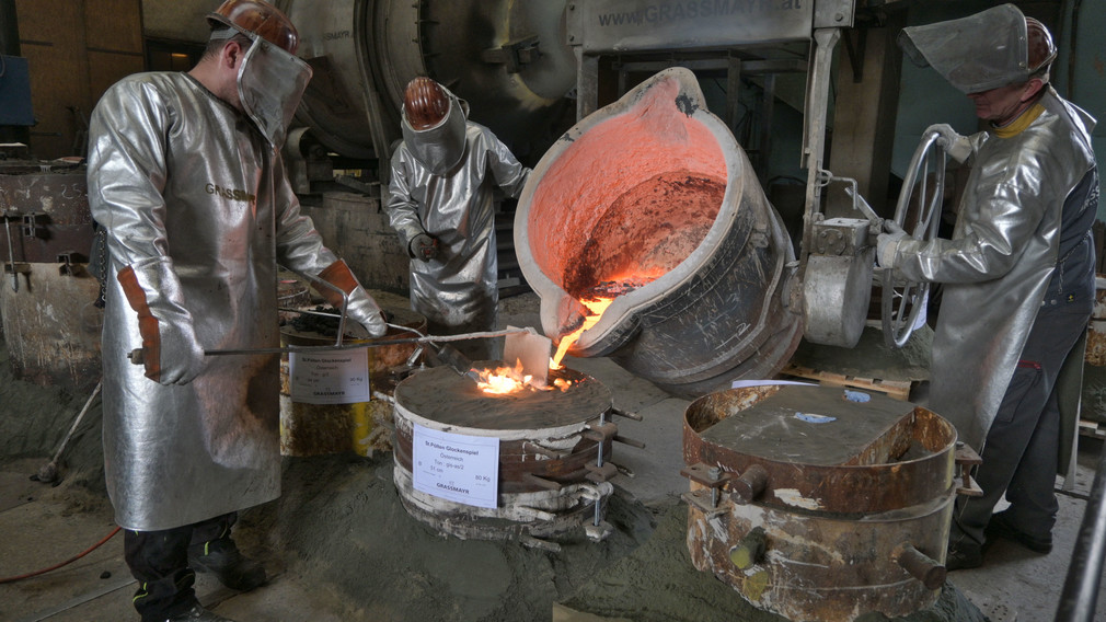 Drei Fachkräfte bei der arbeit: Sie gießen aus geschmolzenem Metall eine Glocke.