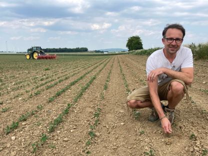 Soja-Feld mit Mann rechts im Bild, der in die Erde mit noch sehr kleinen Sojapflanzen greift. Im Hintergrund links in weiter Entfernung steht ein Traktor. Der Himmel ist blau, durchzogen von weißen Wolken. Foto: Althea Müller