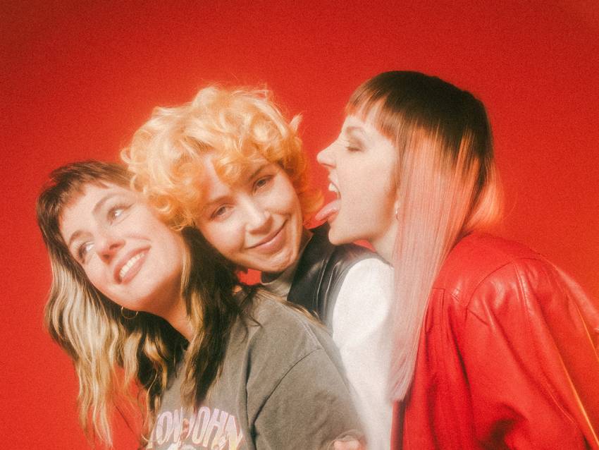 Drei junge Frauen umarmen sich, die rechte Frau streckt die Zunge raus. Der Hintergrund ist rot. Foto: Mala Kolumna