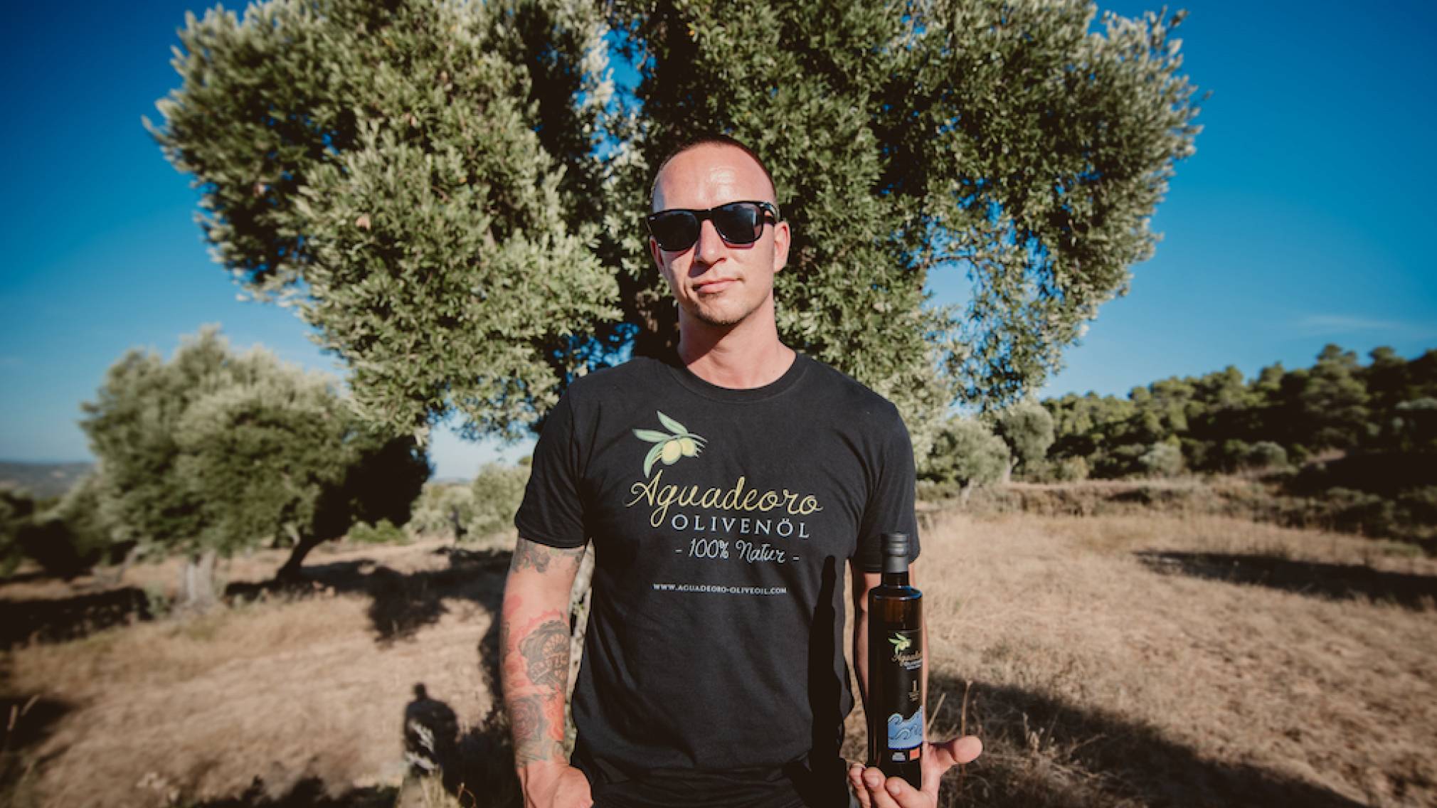In einer spanischen Landschaft steht ein Mann in T-Shirt mit Sonnenbrille vor einem Olivenbaum. In der Hand hält er eine Flasche Olivenöl. Foto: Agrowable/Roberto Bondia