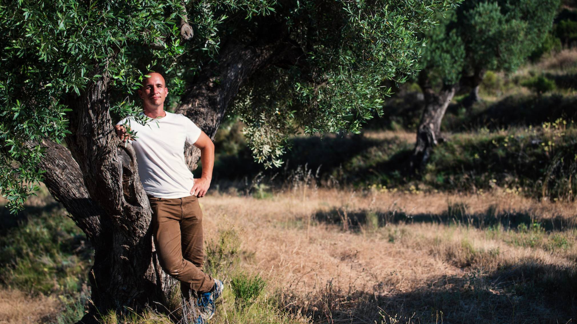 In einer spanischen Landschaft steht ein Mann in T-Shirt mit Sonnenbrille vor einem Olivenbaum. In der Hand hält er eine Flasche Olivenöl. Foto: Agrowable/Roberto Bondia
