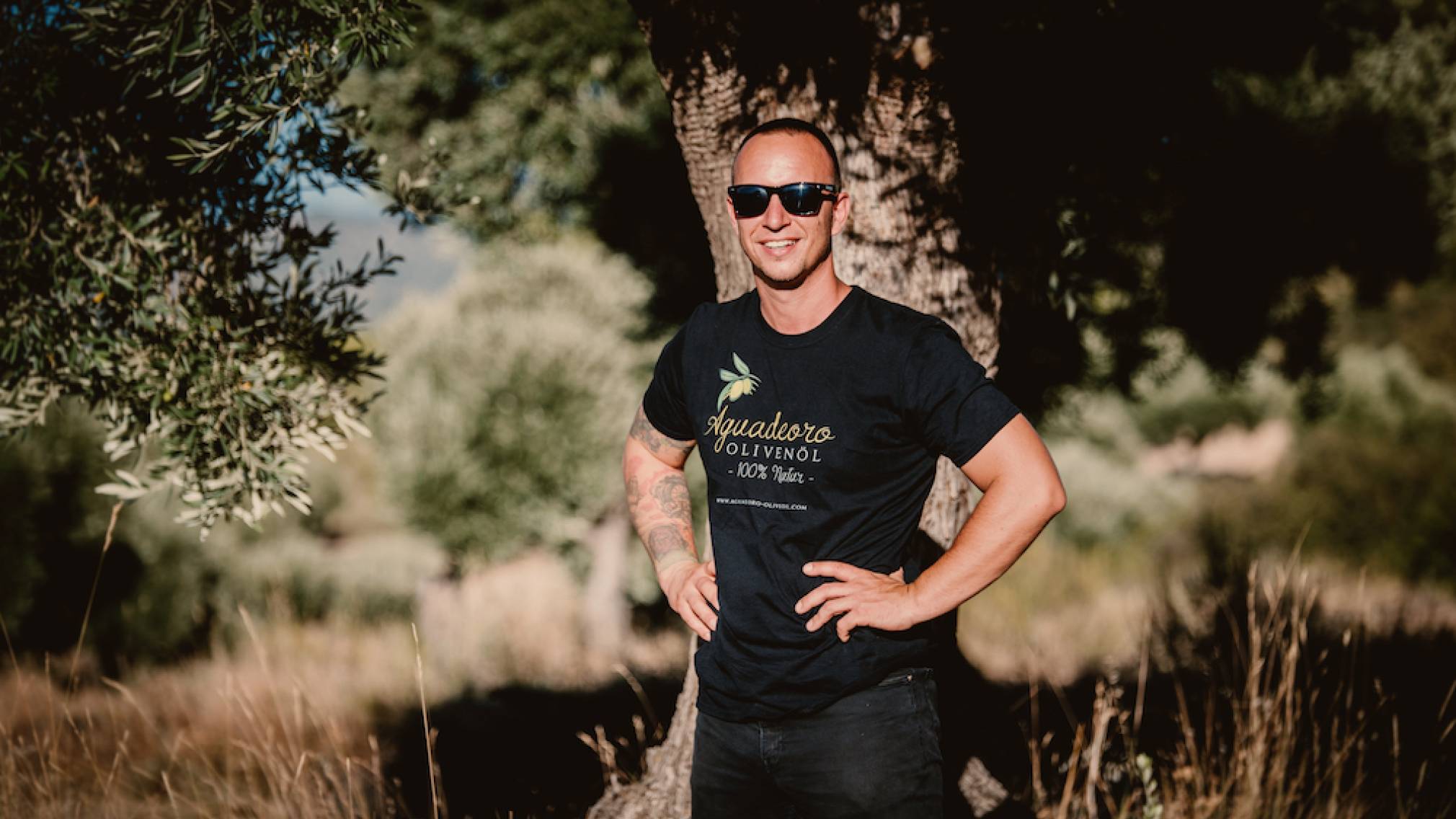 In einer spanischen Landschaft steht ein Mann in T-Shirt mit Sonnenbrille vor einem Olivenbaum. In der Hand hält er eine Flasche Olivenöl. Foto: Agrowable/Roberto Bondia